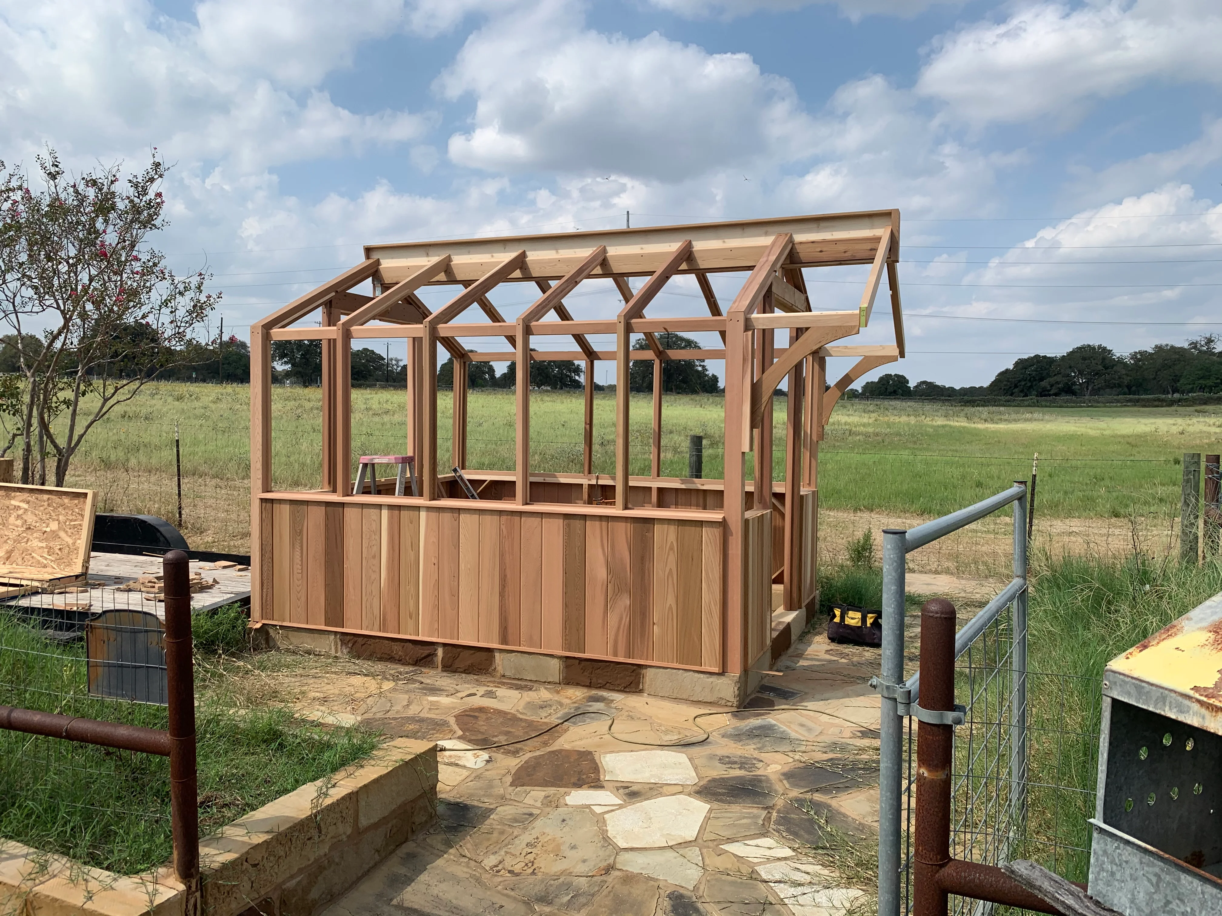 Timber frame pavilion with pergola roof, wood siding, and stone patio foundation in rural setting