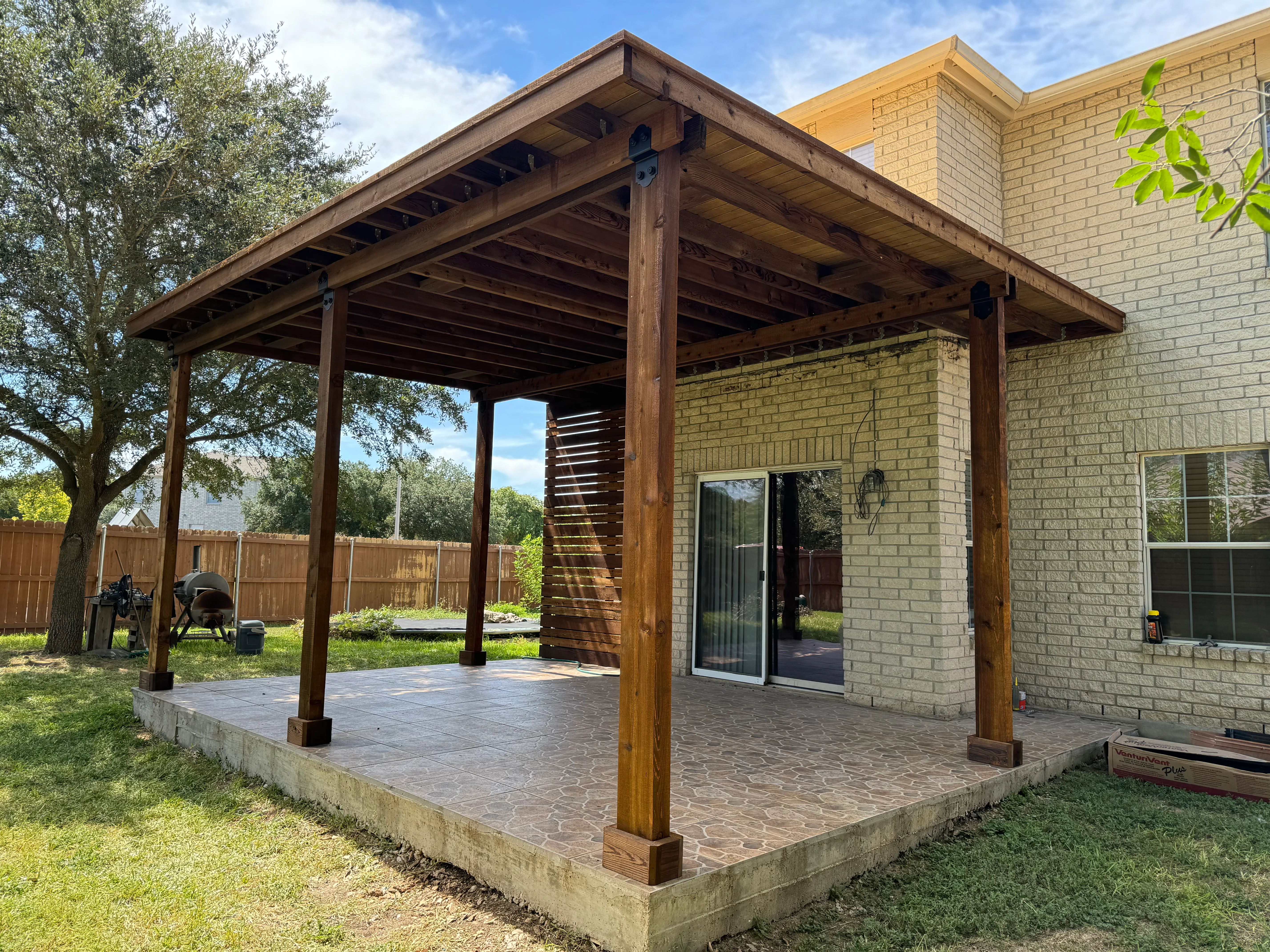 Stained wood pergola with horizontal slat screen over stamped concrete paver patio adjacent to brick home