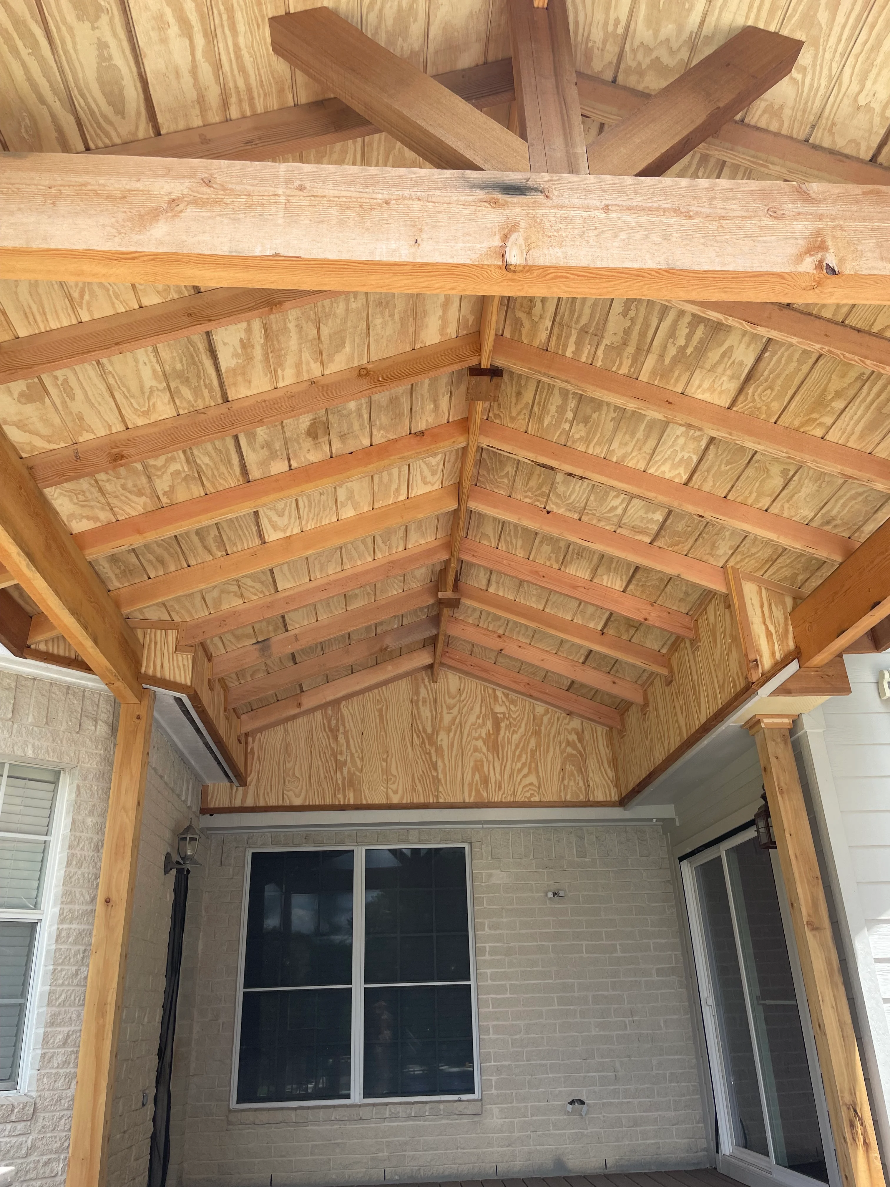 Timber frame patio cover with exposed beams, gabled roof, and OSB sheathing over brick exterior